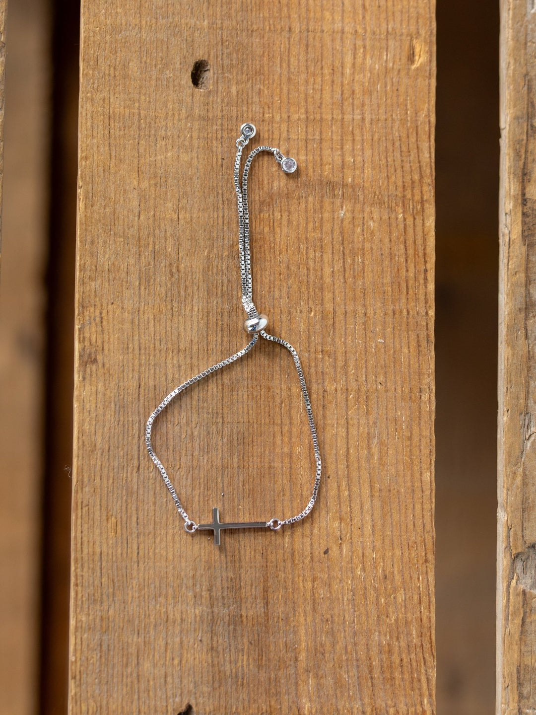 The Bethel Gold/Silver Cross Bracelet by Savannah Sevens western life{&}style, featuring an adjustable 14K gold plated design and a horizontal cross charm, is displayed on a wooden surface.