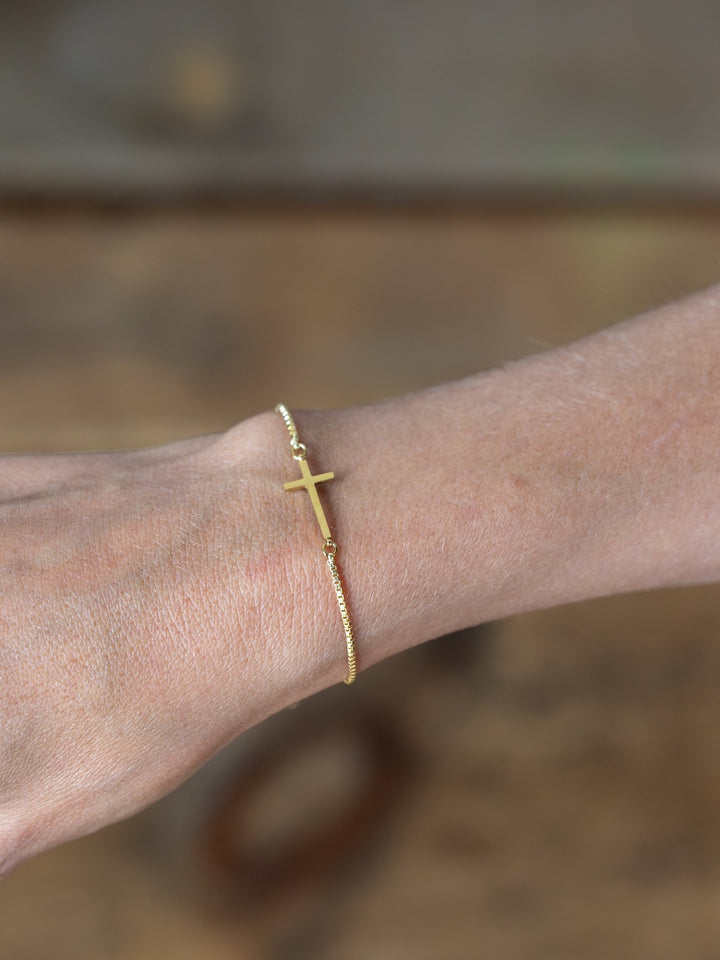 A close-up of a wrist wearing the Bethel Gold Cross Bracelet by Savannah Sevens western life{&}style, set against a blurred brown background.