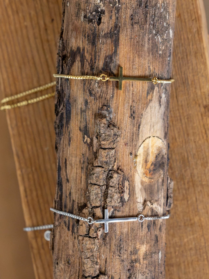The Bethel Gold/Silver Cross Bracelet by Savannah Sevens western life{&}style features adjustable gold-plated and silver options, shown on a rustic branch. Each bracelet adds an elegant touch to any outfit.