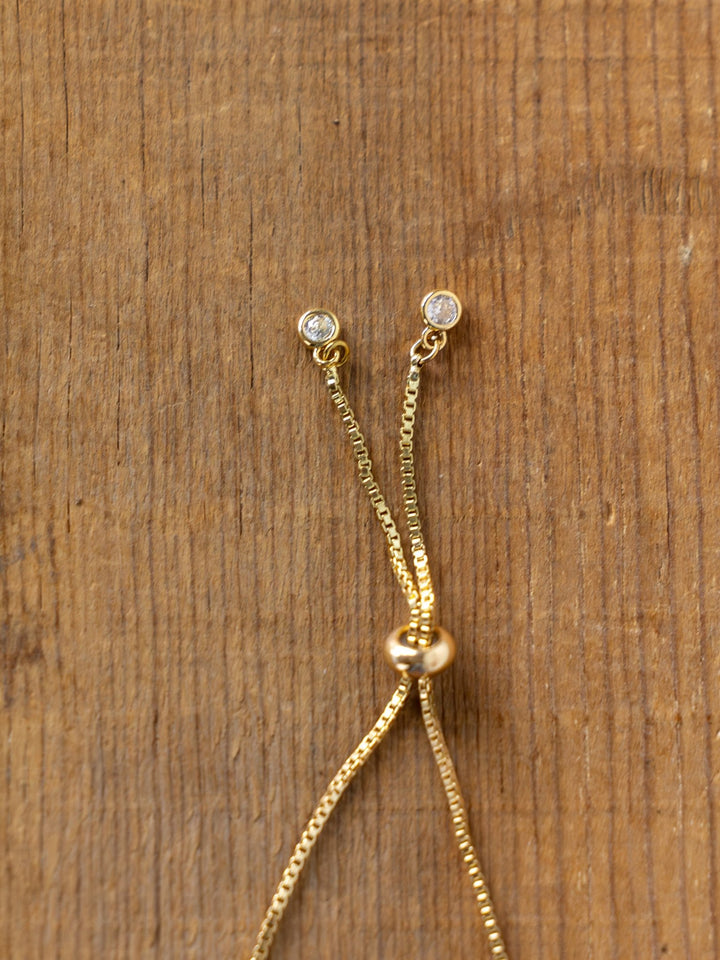 Close-up of Savannah Sevens western life{&}style Bethel Gold/Silver Cross Bracelets featuring adjustable chains with small round clear gemstones and a sliding gold bead, set against a wooden background.