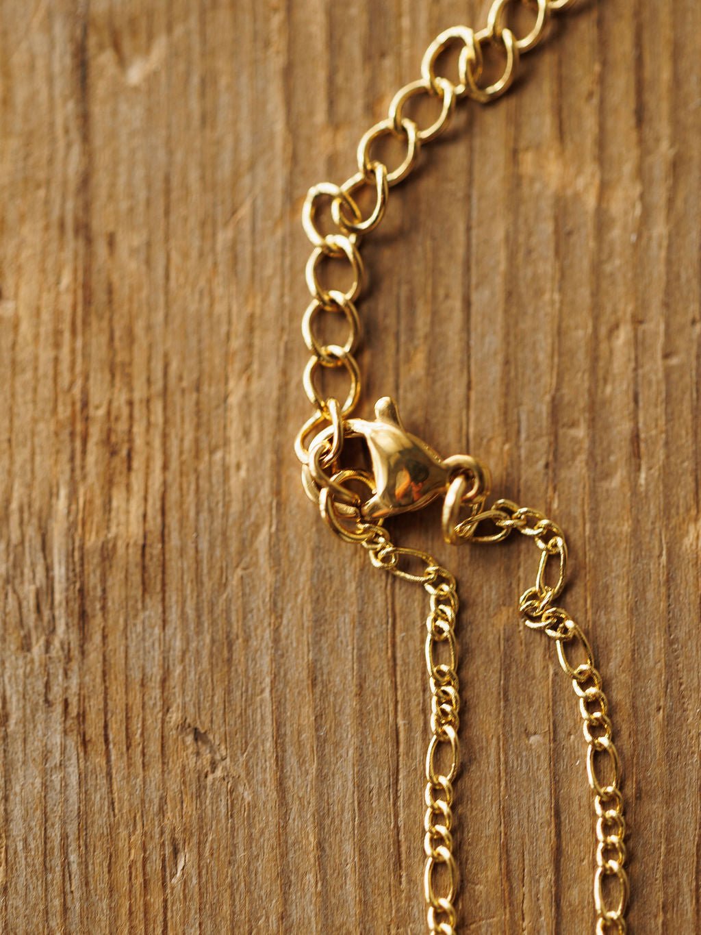 A close-up of the Bethel Necklace by Savannah Sevens western life{&}style, featuring a petite cross and lobster clasp on a textured wood surface. The detailed chain highlights its hypoallergenic design.