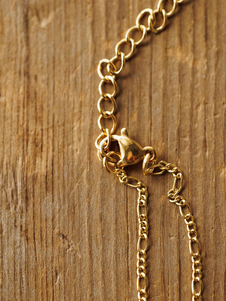A close-up of the Bethel Necklace by Savannah Sevens western life{&}style, featuring a petite cross and lobster clasp on a textured wood surface. The detailed chain highlights its hypoallergenic design.