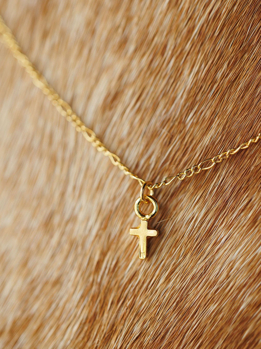The Bethel Necklace by Savannah Sevens western life{&}style features a petite gold cross pendant on a light brown textured surface. This hypoallergenic piece combines comfort with elegant style.