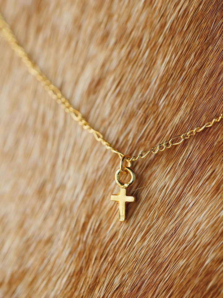 The Bethel Necklace by Savannah Sevens western life{&}style features a petite gold cross pendant on a light brown textured surface. This hypoallergenic piece combines comfort with elegant style.