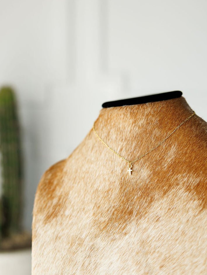 The Bethel Necklace by Savannah Sevens western life{&}style, featuring a petite hypoallergenic gold cross pendant, is shown on a brown and white cowhide mannequin with a blurred cactus in the background.