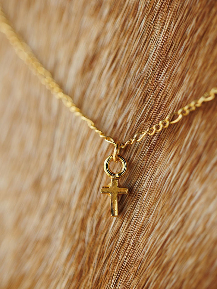 A close-up of the hypoallergenic Bethel Necklace by Savannah Sevens western life{&}style, featuring a petite gold cross pendant resting against light brown cowhide.