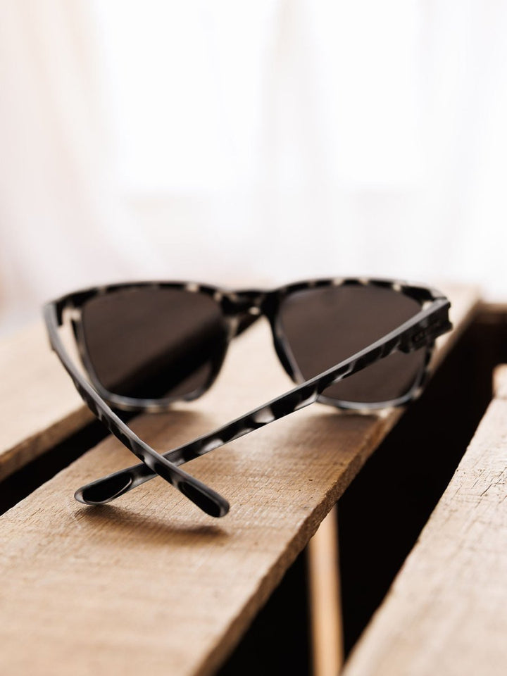 The Bex Adams Sunglasses in Tortoise Gray by Savannah Sevens western life{&}style sit on a wooden slatted surface, highlighted against a softly blurred, light background.