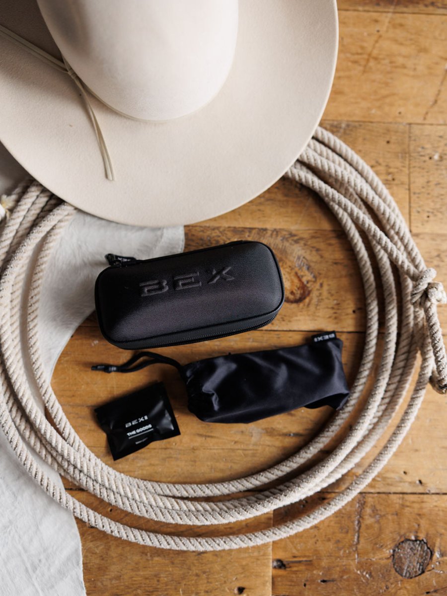 A beige cowboy hat, coiled rope, and a black Savannah Sevens BEX case sit on rustic wood—ideal for carrying Bex Adams Sunglasses {Tortoise Gray}, which feature polarized lenses and UV protection.