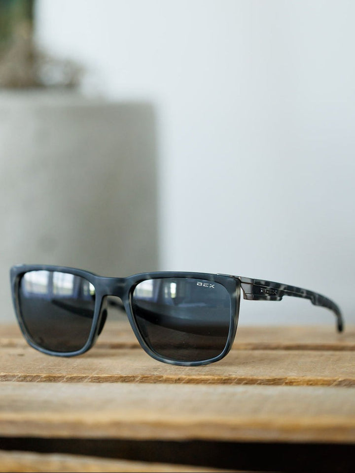 A pair of Bex Adams Sunglasses in Tortoise Gray by Savannah Sevens western life{&}style sits on a wooden surface, with a blurred gray pot in the background.