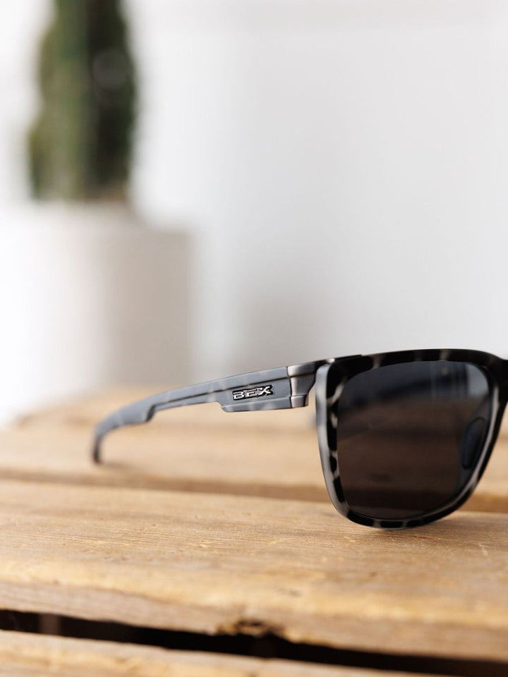 The Bex Adams Sunglasses in Tortoise Gray by Savannah Sevens western life&style, featuring polarized lenses and UV protection, rest on a wooden surface with a blurred cactus and white background in the distance.