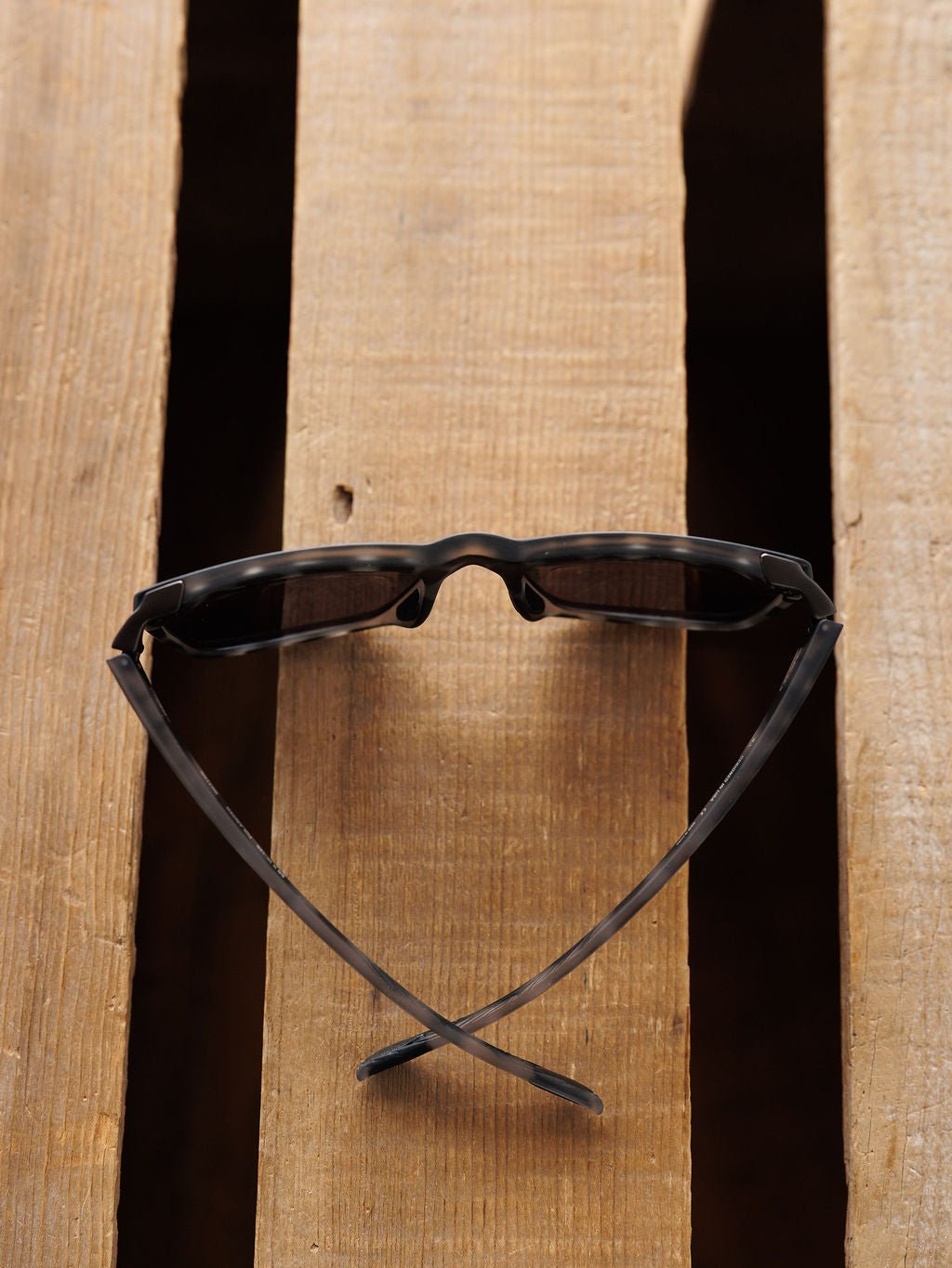 A pair of Bex Adams Sunglasses in Tortoise Gray by Savannah Sevens western life{&}style rests upside down on a wooden surface, with arms extended and crossed to resemble a smiley face.