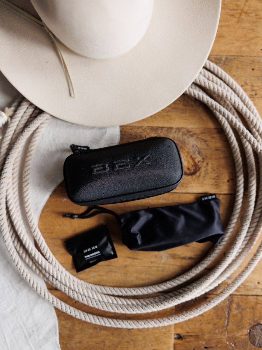 A beige cowboy hat, coiled rope, and a black BEX case with lens cloth pouch and wipe rest on wood, capturing western flair—ideal for storing Savannah Sevens western life&style Bex Fynnland XL Sunglasses {Black} with UV protection.
