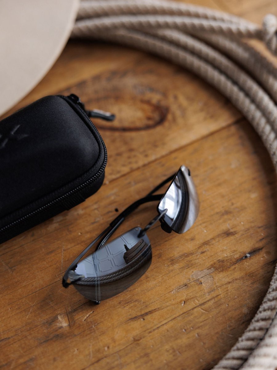 The Bex Fynnland XL Sunglasses in black by Savannah Sevens western life&style rest beside a black zippered case on a wooden surface, with coiled rope in the background.