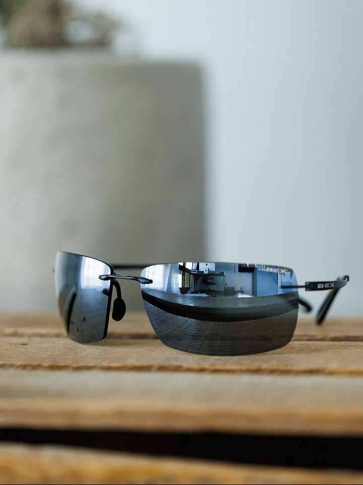The Bex Fynnland XL Sunglasses in black by Savannah Sevens western life{&}style rest on a wooden surface, their polarized lenses reflecting an indoor room, with a blurred gray planter visible in the background.