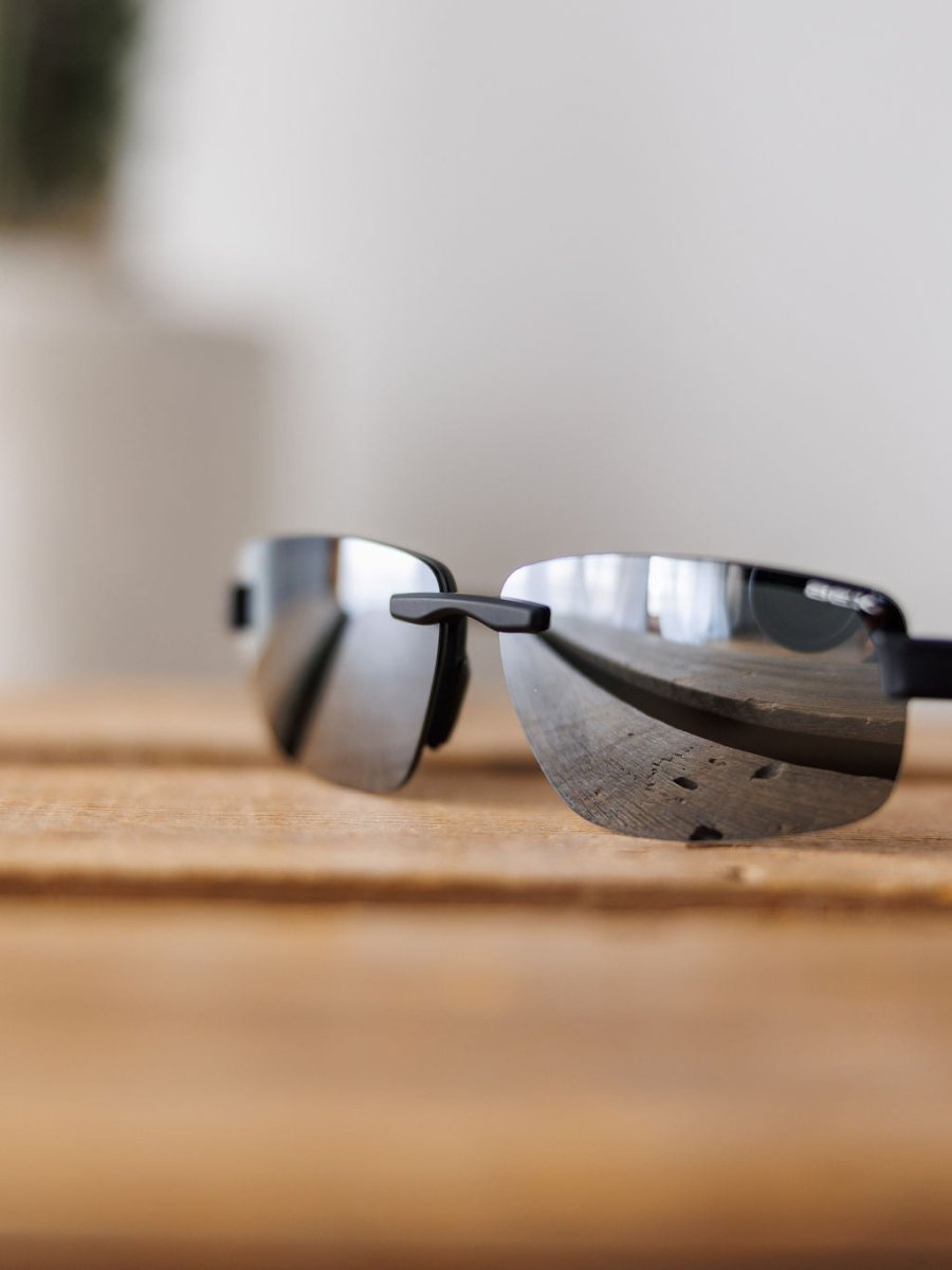 A close-up of Bex Harris Sunglasses {Black} by Savannah Sevens western life{&}style, featuring sleek polarized lenses resting on wood and softly reflecting their surroundings with a mirrored finish against a blurred backdrop.