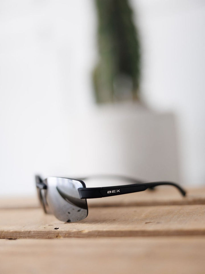 A pair of Bex Harris Sunglasses in black from Savannah Sevens western life{&}style rests on a wooden surface, with a blurred white pot and green plant in the background.