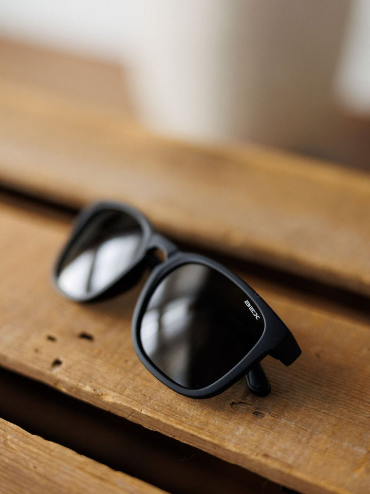 The Bex Zion Sunglasses in black by Savannah Sevens western life&style, featuring polarized scratch-resistant lenses and a matte frame, rest at an angle on a wooden surface.