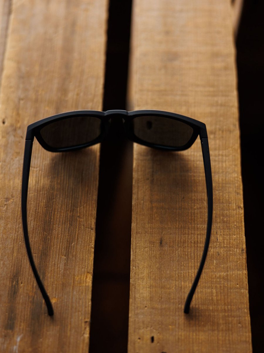 A pair of Bex Zion Sunglasses in black by Savannah Sevens western life{&}style, featuring polarized, scratch-resistant lenses, rests upside down on a wooden surface with wide plank gaps.