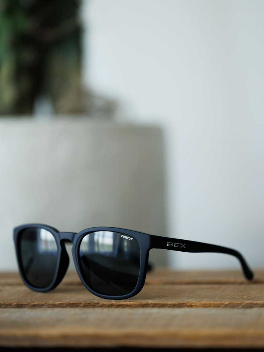 Bex Zion Sunglasses in black by Savannah Sevens western life{&}style feature dark, scratch-resistant lenses with UVA/UVB protection and “BEX” branding on the arm, shown resting on wood with a blurred background.