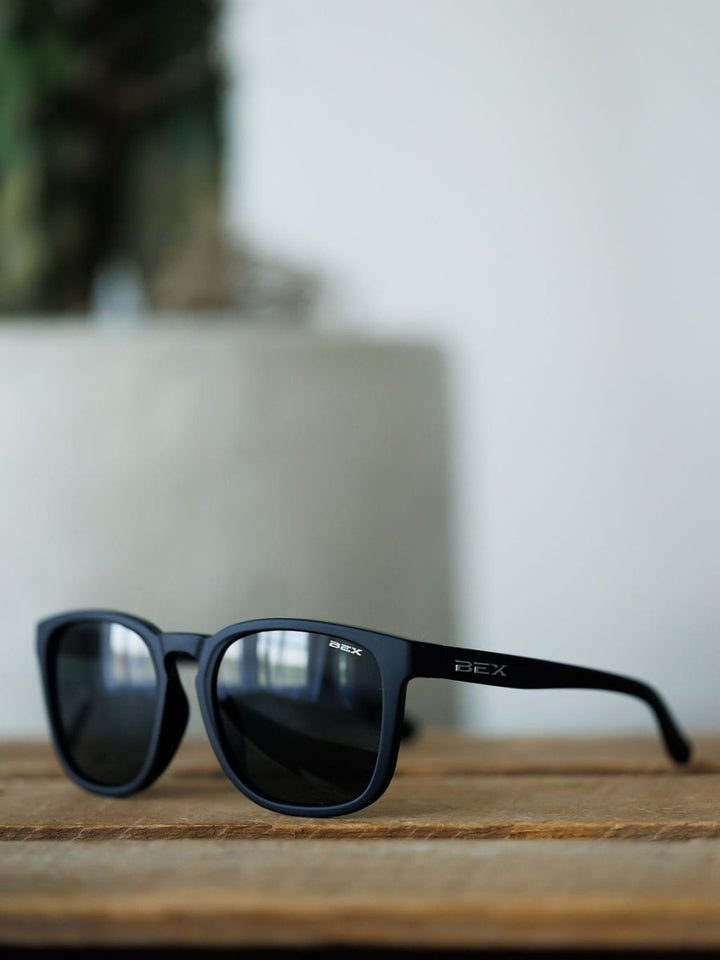 Bex Zion Sunglasses in black by Savannah Sevens western life{&}style feature dark, scratch-resistant lenses with UVA/UVB protection and “BEX” branding on the arm, shown resting on wood with a blurred background.