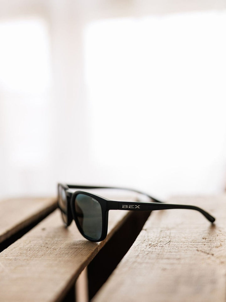 A pair of Bex Zion Sunglasses in black by Savannah Sevens western life&style, featuring polarized, scratch-resistant lenses, rests on a wooden surface against a bright, blurred background.