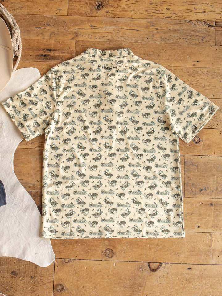A short-sleeved, cream-colored Big Bass Cinch Men's Graphic Polo by Savannah Sevens western life{&}style, featuring a pattern of small fish illustrations, lies flat on a wooden floor. The moisture-wicking bass fishing shirt is complemented by the edges of a cowhide and rope.