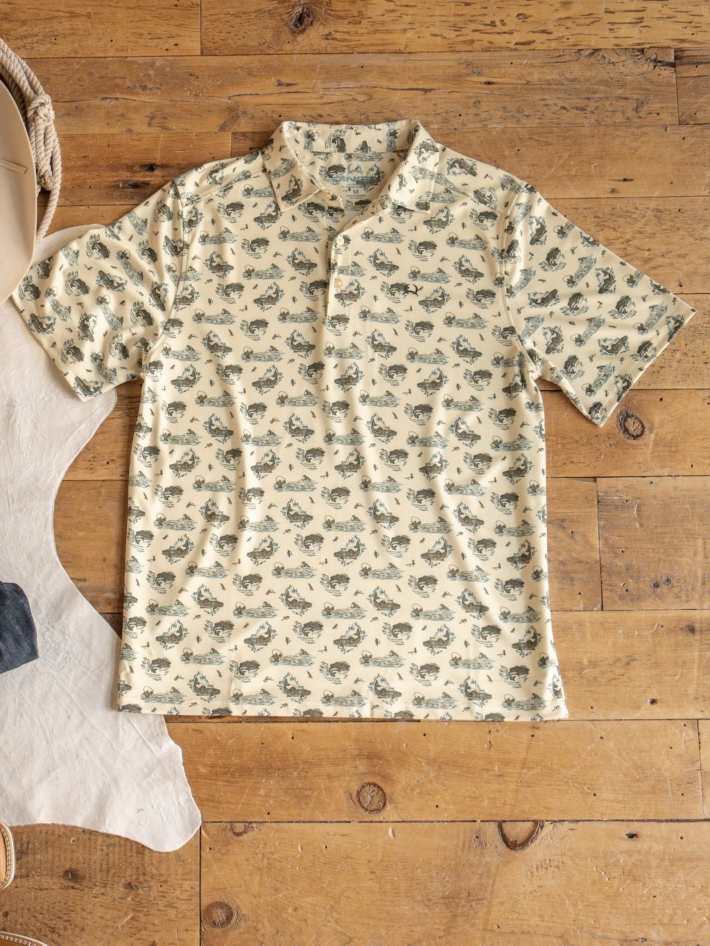The Big Bass Cinch Men's Graphic Polo by Savannah Sevens western life{&}style—a beige short-sleeve ArenaFlex shirt with an all-over fish print—is laid flat on a wooden floor beside part of a white rug and some denim fabric. This polo features moisture-wicking fabric to keep you comfortable.