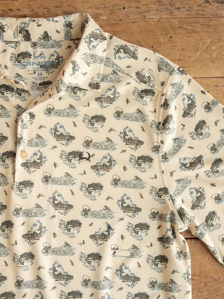 A Big Bass Cinch Men's Graphic Polo by Savannah Sevens western life{&}style, featuring a beige color and a pattern of fish, water, and reeds, lies on a wooden surface. Three buttons are visible on the shirt’s placket.