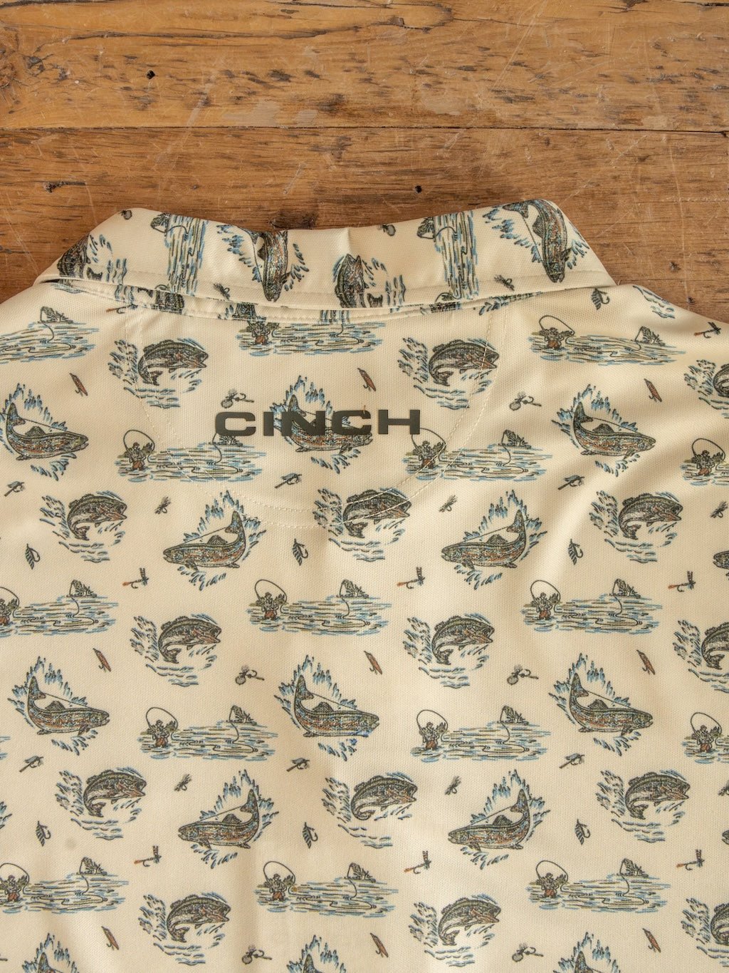 Introducing the Big Bass Cinch Men's Graphic Polo by Savannah Sevens western life{&}style—a beige ArenaFlex polo adorned with a subtle green nature print of trees, water, and canoes. The word "CINCH" stands out in large gray letters across the upper back. Laid on a wooden surface, this shirt offers moisture-wicking comfort designed for all-day wear.