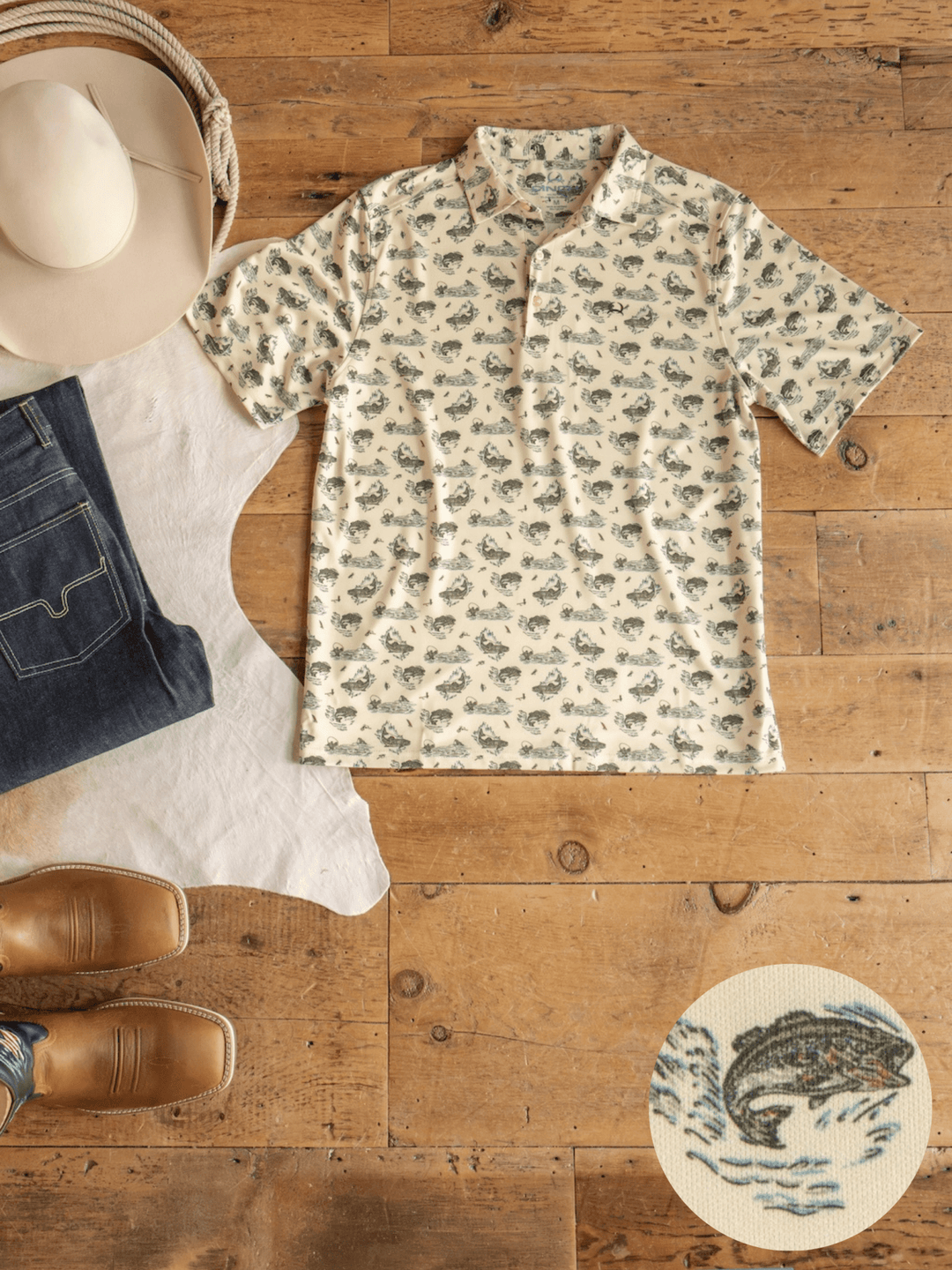 A cream Big Bass Cinch Men's Graphic Polo from Savannah Sevens western life{&}style, featuring a repeating bass fish print, is displayed on a wooden floor alongside jeans, a cowboy hat, brown boots, and a white cowhide rug. This moisture-wicking polo offers comfort and performance for bass fishing. A close-up provides a detailed look at the fish pattern.