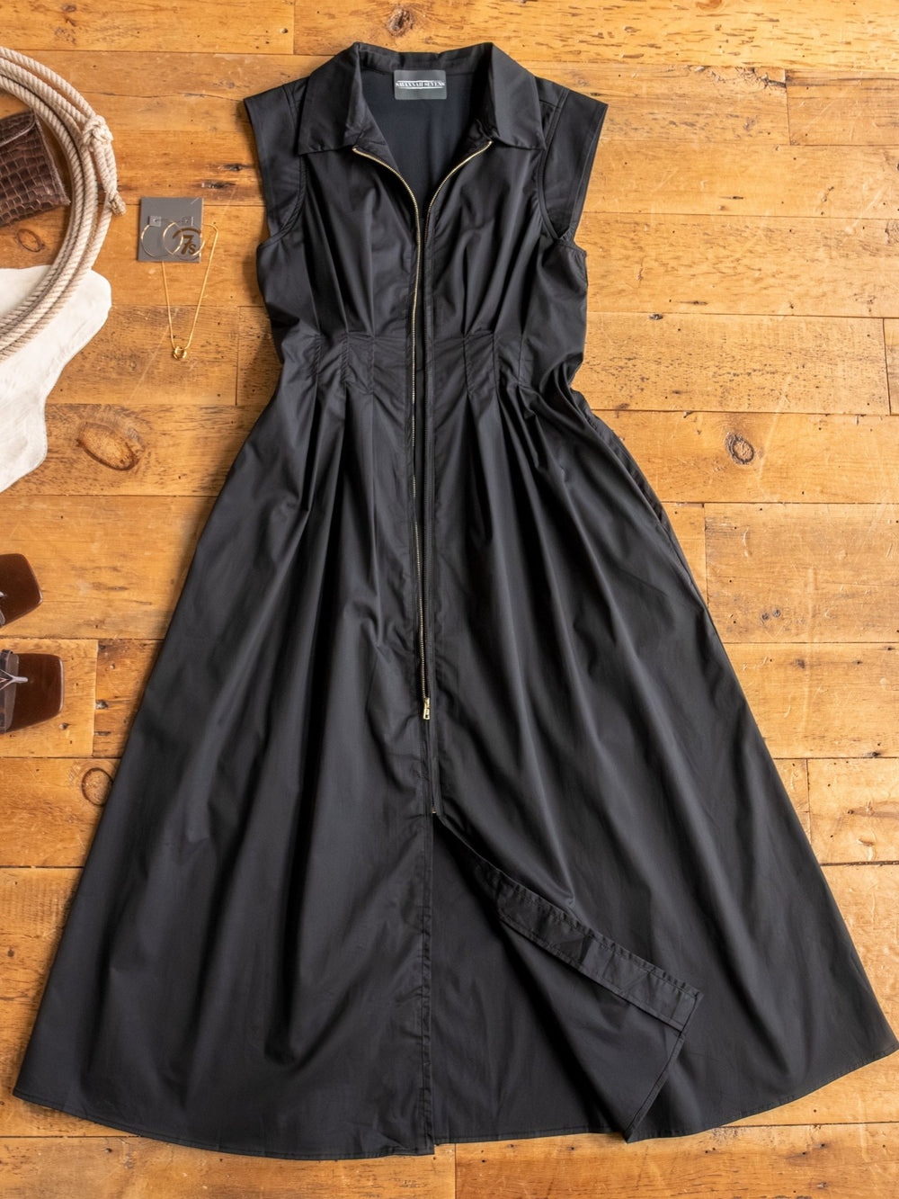 The Biloxi by Savannah Sevens western life{&}style—a black cap sleeve midi dress with front zipper and flared skirt—is laid flat on a wooden floor, with a necklace, card, and small brown items arranged beside it.