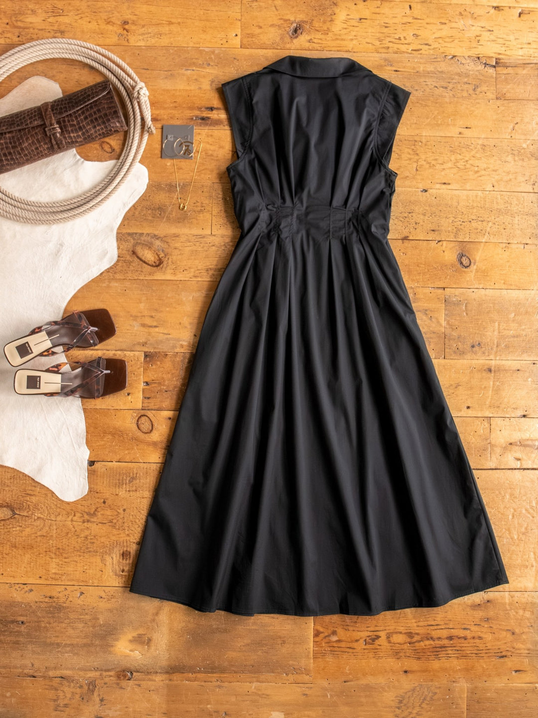 The Biloxi dress by Savannah Sevens western life{&}style, a black cotton blend piece, is laid flat on a wooden floor with a cowhide rug, heeled sandals, and a small jewelry box with earrings nearby.