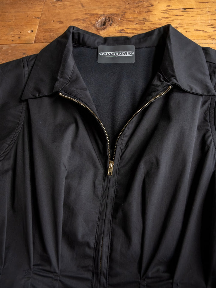 A close-up of The Biloxi black cap sleeve midi dress with a front zipper, laid flat on wood. The inside label shows the brand Savannah Sevens western life{&}style.