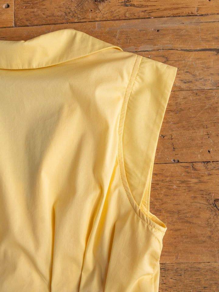 A close-up of the Biloxi Butter Zip Front Dress by Savannah Sevens western life{&}style shows its butter yellow cotton blend, gathered fabric, and clean seams. The stylish midi dress is laid on rustic wood with only its upper portion visible.