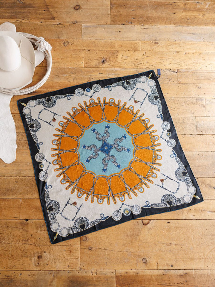 The Bits Wild Rag {Rust} by Savannah Sevens western life{&}style, a vibrant jacquard silk scarf with blue, orange, and white geometric patterns, is displayed flat on a wooden floor beside a woven-band white hat and white scarf.