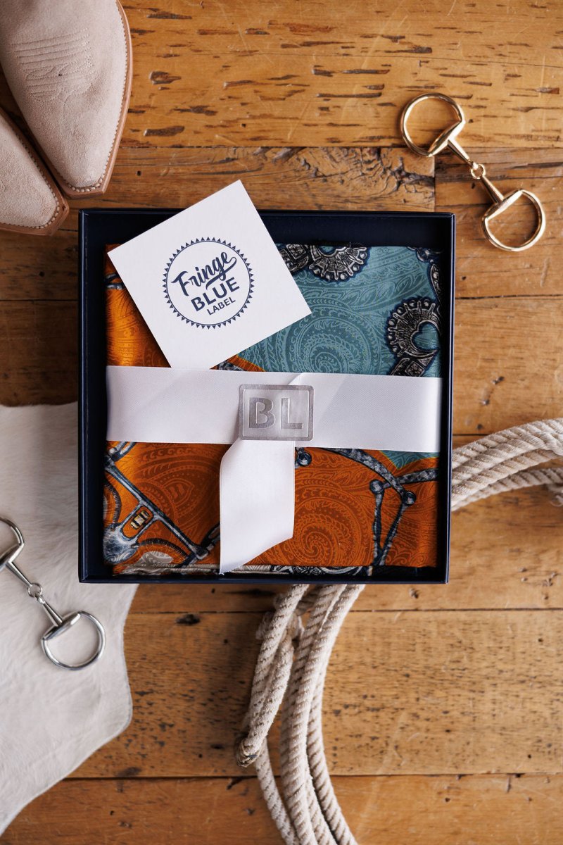 The Bits Wild Rag {Rust} by Savannah Sevens western life{&}style, a vibrant jacquard silk scarf with intricate patterns, comes neatly boxed and ribbon-tied with a silver BL logo, accompanied by a Fringe Blue Label card and luxe accessories.