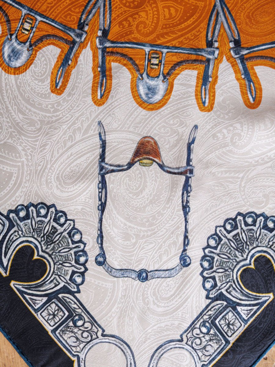 A close-up of the Bits Wild Rag {Rust} by Savannah Sevens western life{&}style, a jacquard silk scarf with intricate orange, white, navy blue, and gray designs featuring bridles, paisley patterns, and ornamental motifs.