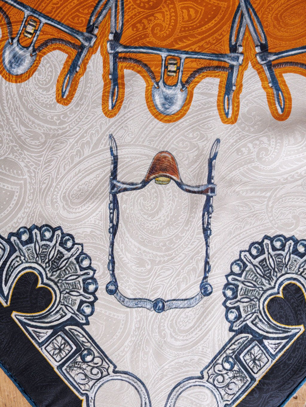 A close-up of the Bits Wild Rag {Rust} by Savannah Sevens western life{&}style, a jacquard silk scarf with intricate orange, white, navy blue, and gray designs featuring bridles, paisley patterns, and ornamental motifs.