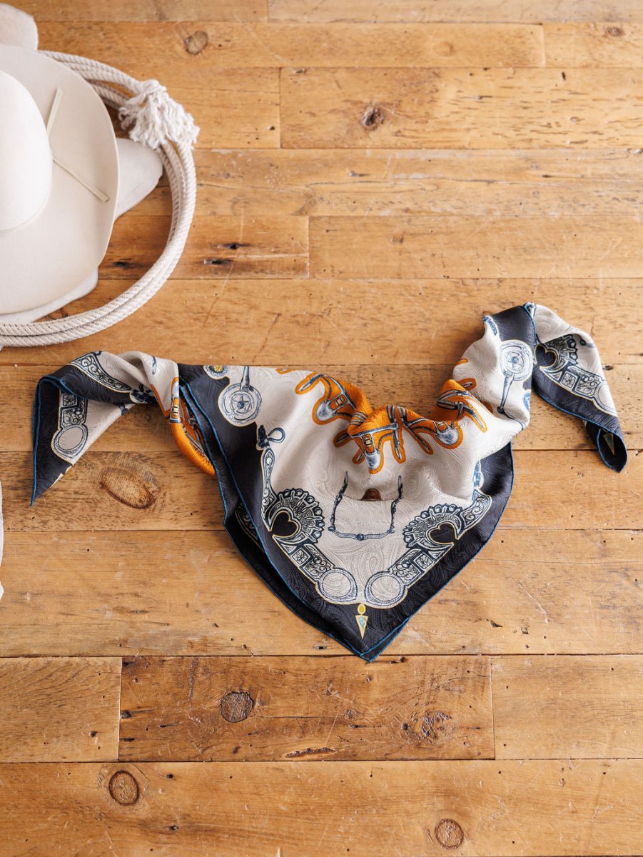 The Bits Wild Rag {Rust} by Savannah Sevens western life{&}style, a patterned paisley silk scarf in black, white, orange, and blue, is folded on wood with a white cowboy hat and coiled rope partly visible in the top left.