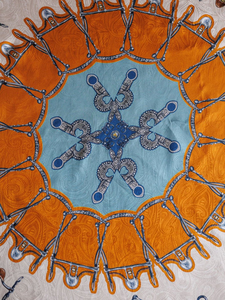 The Bits Wild Rag {Rust} by Savannah Sevens western life{&}style features an ornate circular motif with blue and orange accents, looping designs, and a textured beige background, reminiscent of a jacquard silk scarf or decorative mandala.