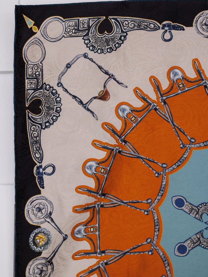 A close-up of the Bits Wild Rag {Rust} by Savannah Sevens western life{&}style, showing ornate jacquard silk with metallic motifs, orange and blue accents, and detailed black, white, and gold symmetrical borders.