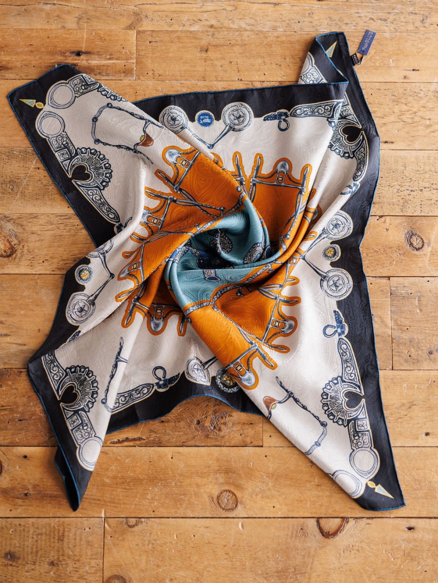 The Bits Wild Rag {Rust} by Savannah Sevens western life{&}style features equestrian-inspired reins, buckles, and rope motifs in black, cream, orange, and teal, artfully displayed on a rustic wooden surface.