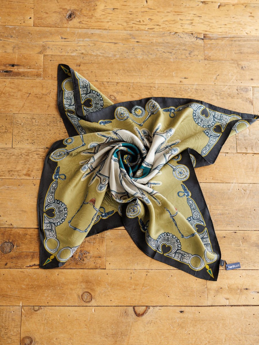 The Bits Wild Rag {Teal} by Savannah Sevens western life{&}style is a jacquard silk scarf with intricate designs, artfully displayed on a rustic wooden floor and finished with a small sewn corner tag.