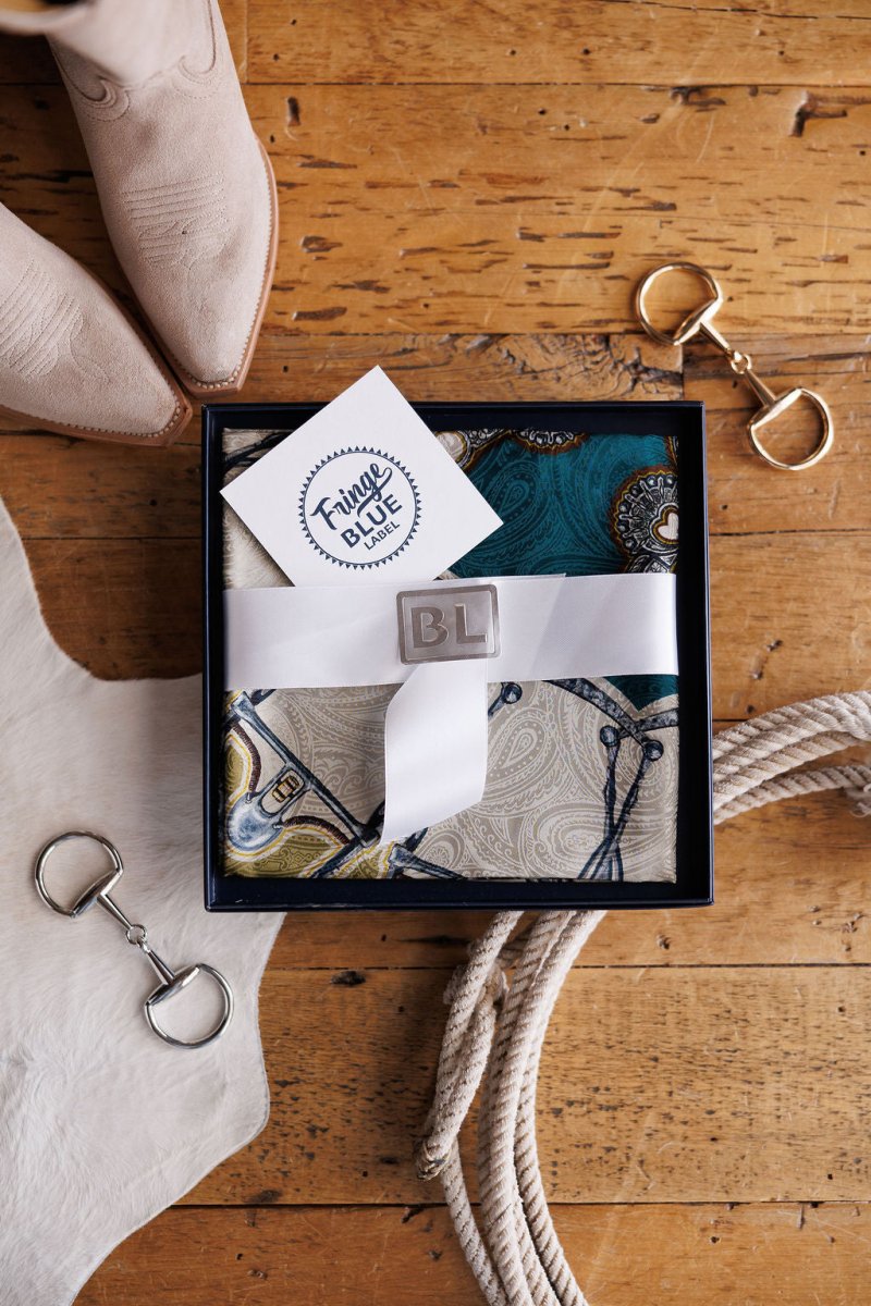 A gift box with a white ribbon and BL engraved on a silver buckle holds the Bits Wild Rag {Teal} scarf from Savannah Sevens western life{&}style. On the wooden floor are beige boots, horse tack, rope, and a Fringe Blue note card nearby.