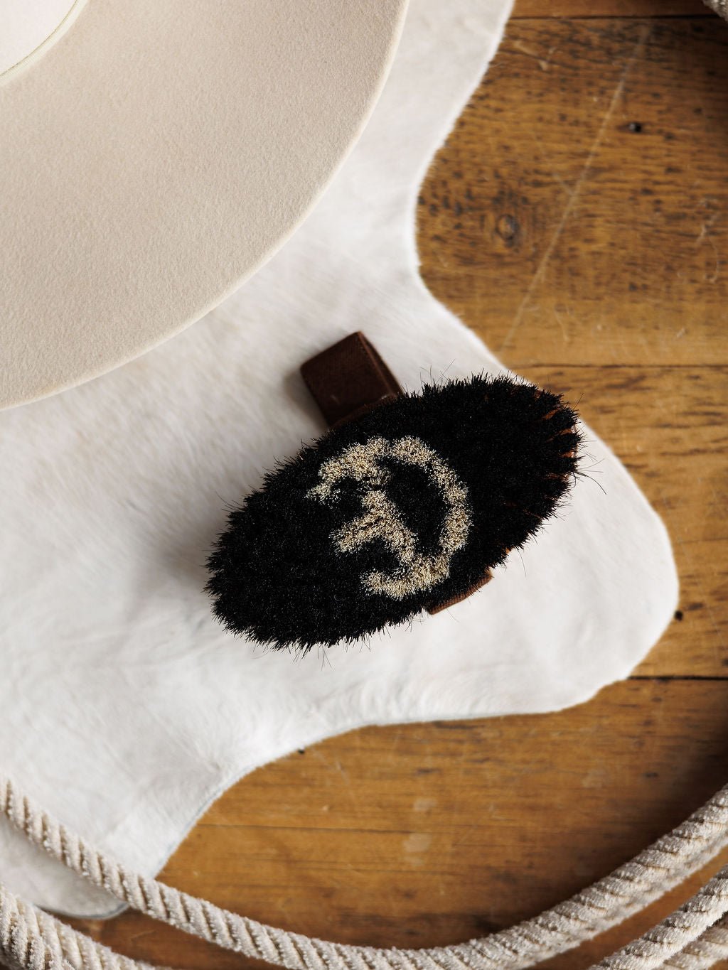 The Savannah Sevens western life{&}style Blanket Brush is a black oval wool brush with pale spiral bristles. It removes pilling from luxury blankets and rests on white hide near a beige hat and coiled rope on wood.