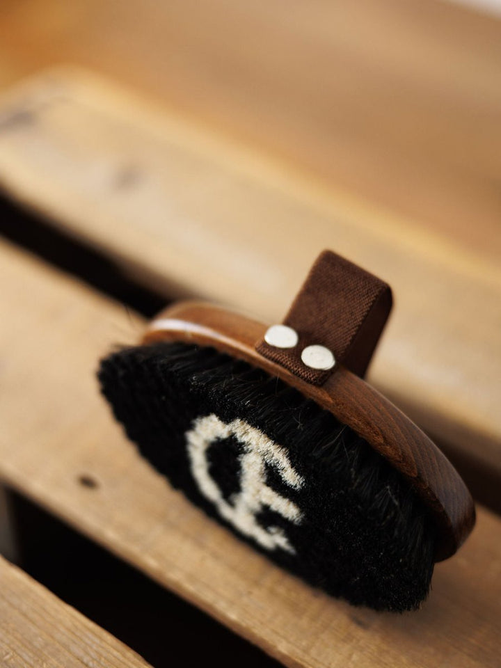 A close-up of the Savannah Sevens western life{&}style Blanket Brush shows its wooden body, dark bristles with a white logo, and leather strap with metal rivets—ideal for luxury blankets and removing pilling.