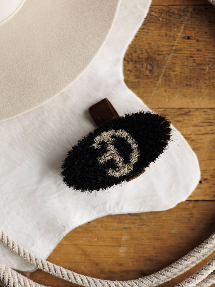 The Savannah Sevens western life{&}style Blanket Brush is a black oval wool brush with pale spiral bristles. It removes pilling from luxury blankets and rests on white hide near a beige hat and coiled rope on wood.