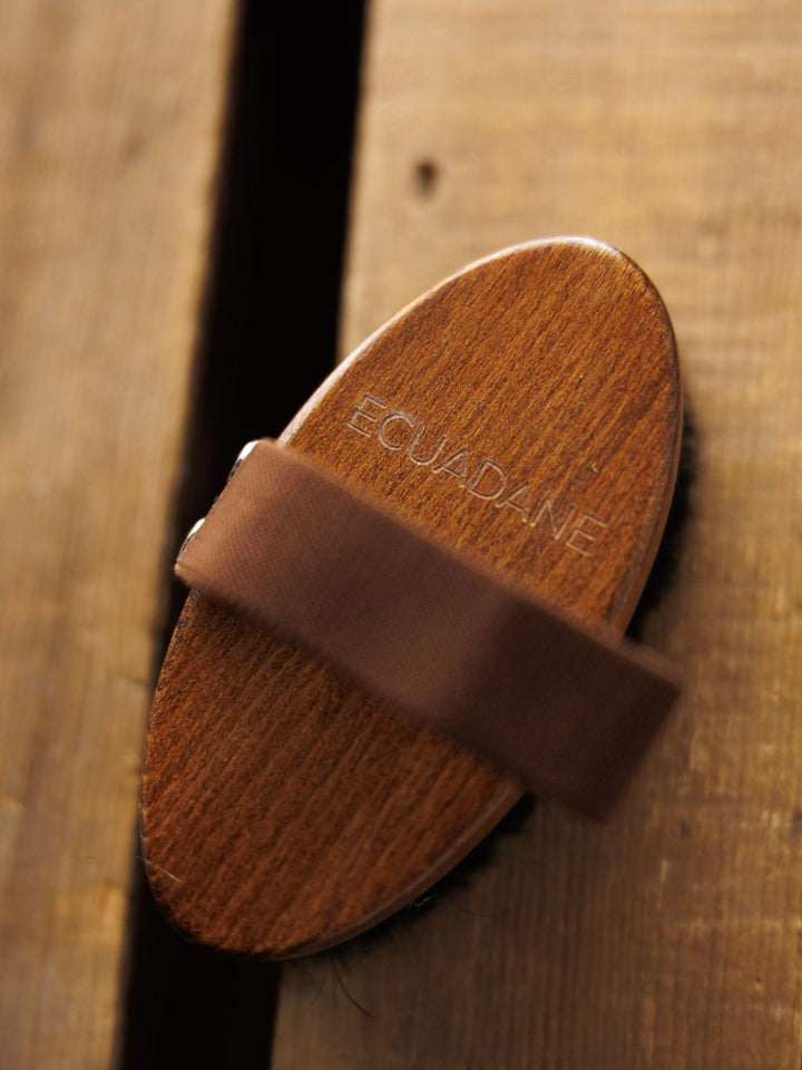 The Blanket Brush by Savannah Sevens western life{&}style, with a wooden body and brown strap, rests on rustic wood. It removes pilling to help maintain the look and feel of your luxury blankets.