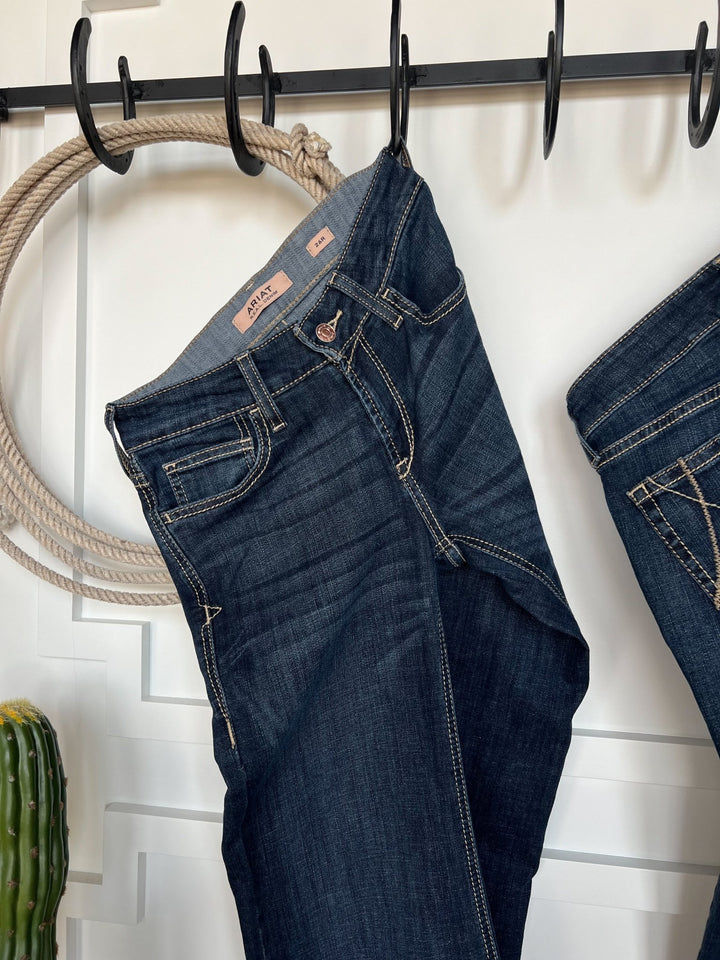 The Blayze Perfect Rise Trouser Jean *Exclusive* by Savannah Sevens western life{&}style in medium dark wash hangs from black hooks on a white wall, with a coiled rope and small cactus in the background.