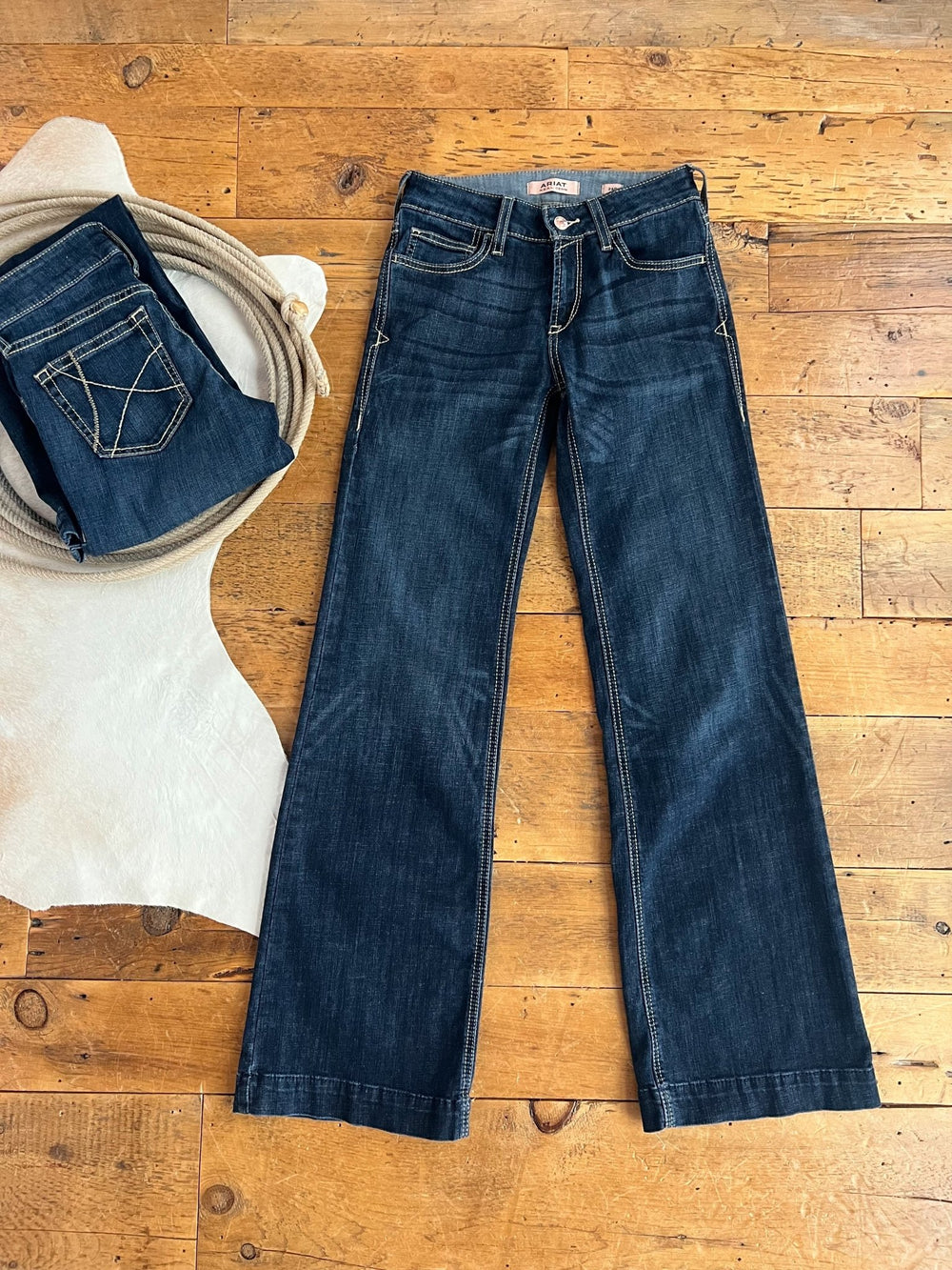 The Blayze Perfect Rise Trouser Jean *Exclusive* by Savannah Sevens western life{&}style in medium dark wash is displayed flat on a wooden floor beside a wicker basket with two folded jeans and a white cowhide rug in the corner.